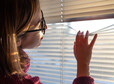 A woman with glasses looks through the blinds at the early morning sunlight.