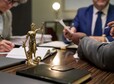 Closeup of Lady Justice statue standing on table with multiethnic group of middle aged men discussing legal documents in office setting, hands holding pens and papers, focus on law theme