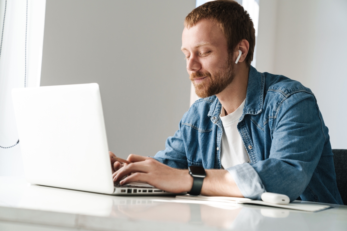 Photo of pleased ginger man using wireless earphones while working with laptop at table indoors