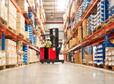Worker in forklift-truck loading packed goods in huge distribution warehouse with high shelves.
