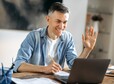 Cheerful handsome successful mature  Caucasian businessman, freelancer or manager in stylish clothes, using laptop, talking to colleagues or clients on a video call, greeting with hand gesture, smiles