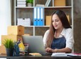 Happy young Asian business owner woman prepare parcel box and check online orders of product for deliver to customer on laptop computer. Shopping Online concept.